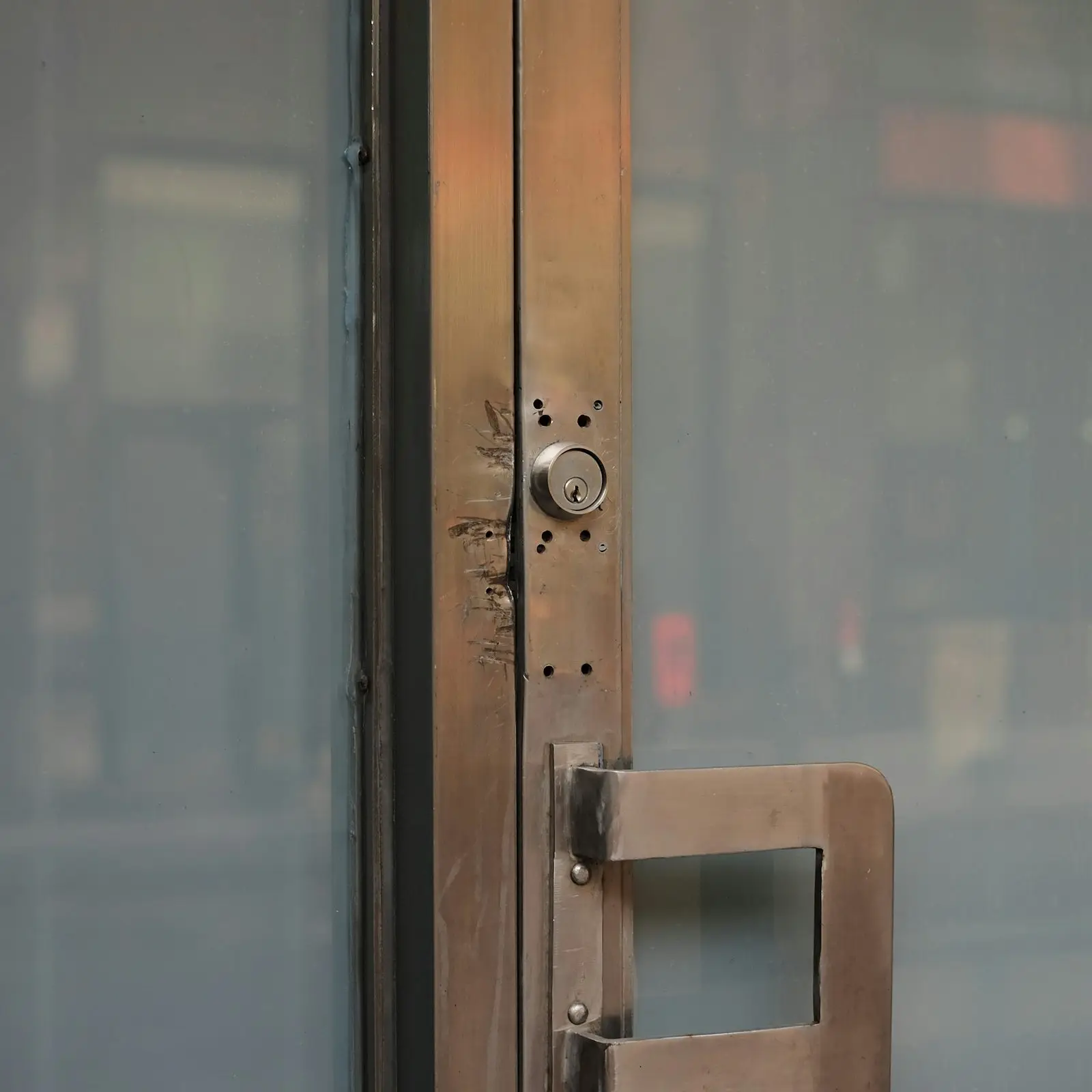 A worn lock cylinder and pull handle on a glass door stile